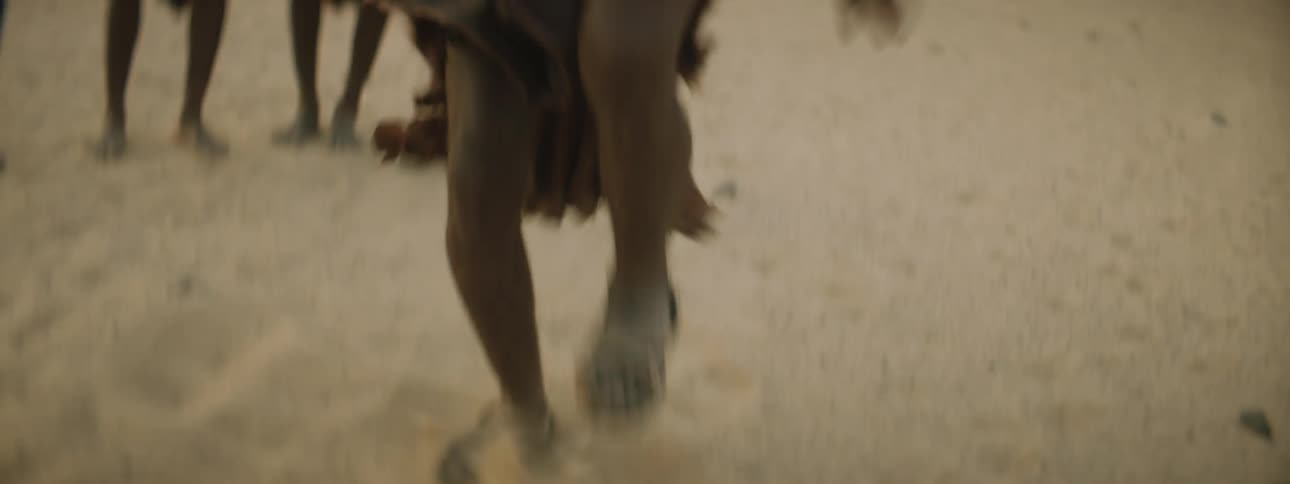 A middle aged man in tribal clothing looks into the camera as he dances in the desert while a group of younger men watch him and clap