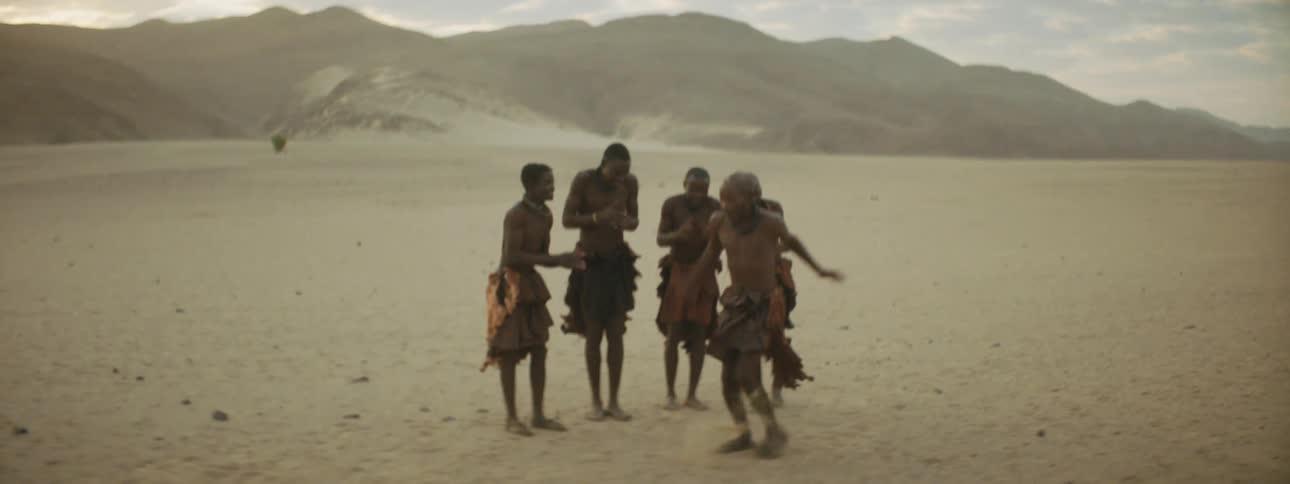 A man does a tribal dance in a desert as other men clap for him