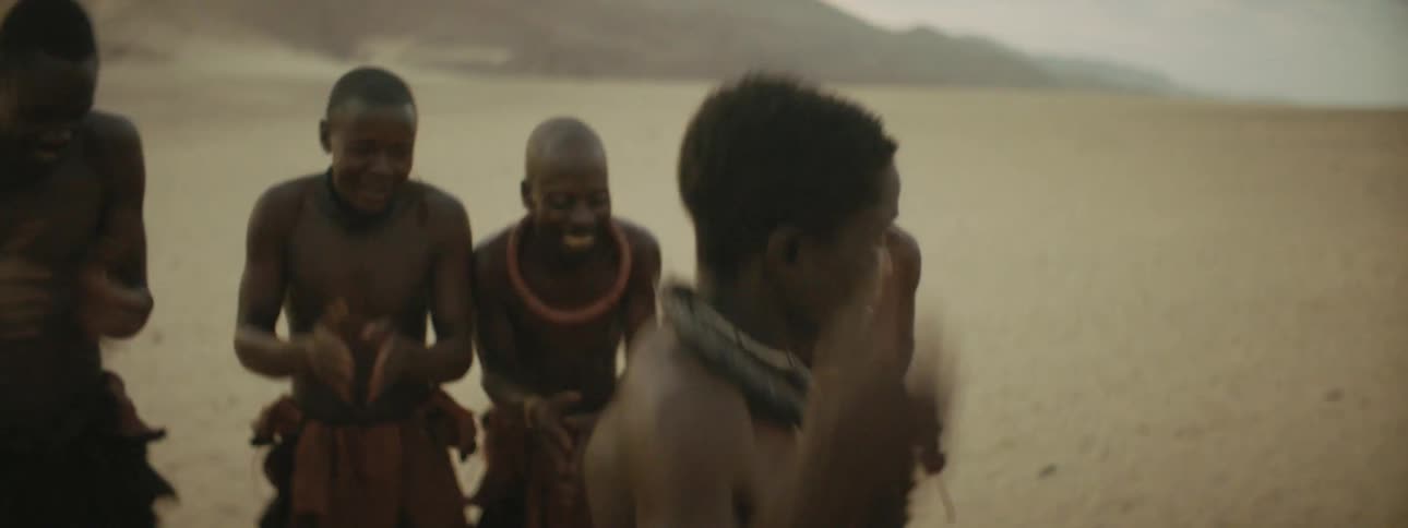 A young man in tribal wear dances in a desert as a group of similarly dressed men watch and clap