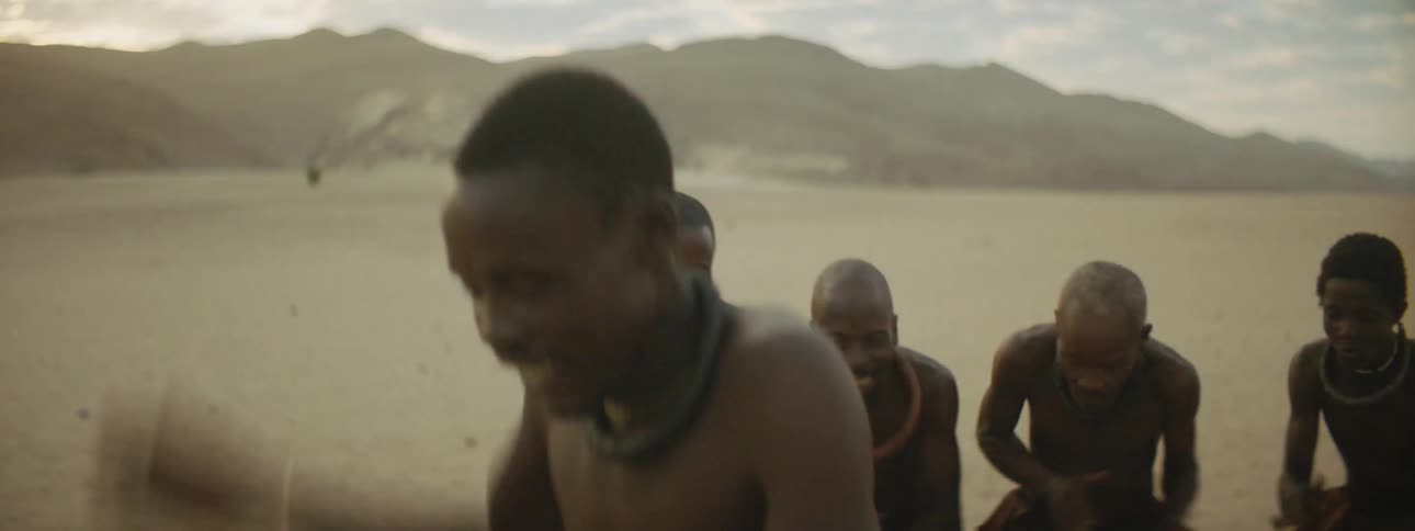 Man dances on sand near hills while friends clap and laugh