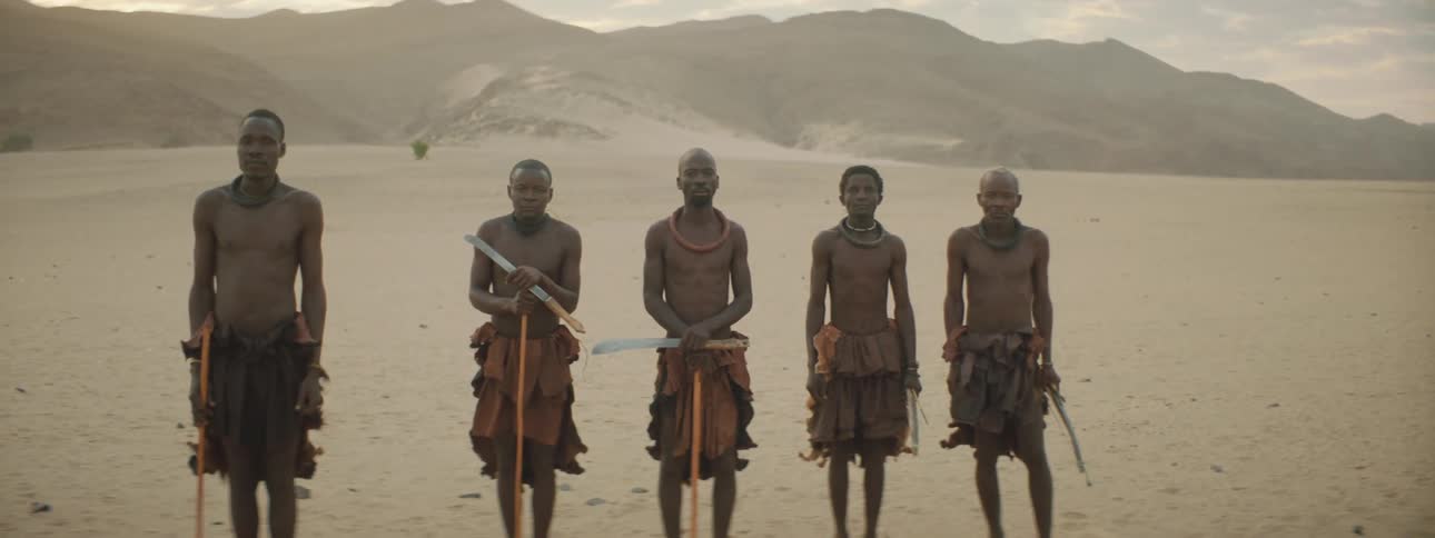 Shirtless men stand holding primitive weapons in the desert near a mountain range
