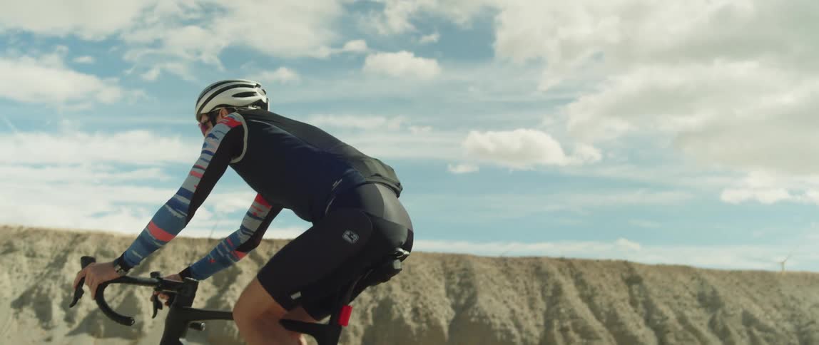 A man wearing a bicycle helmet and sunglasses is riding a bike on a road near mountains under a cloudy sky
