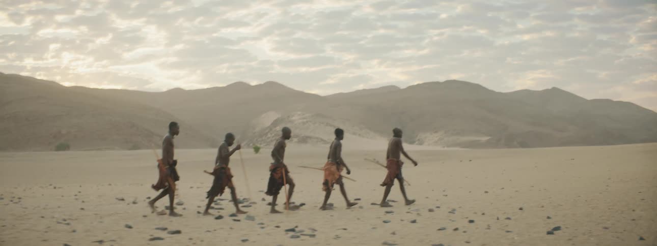Small group of men wearing traditional garb walk across sand near misty mountains carrying bows