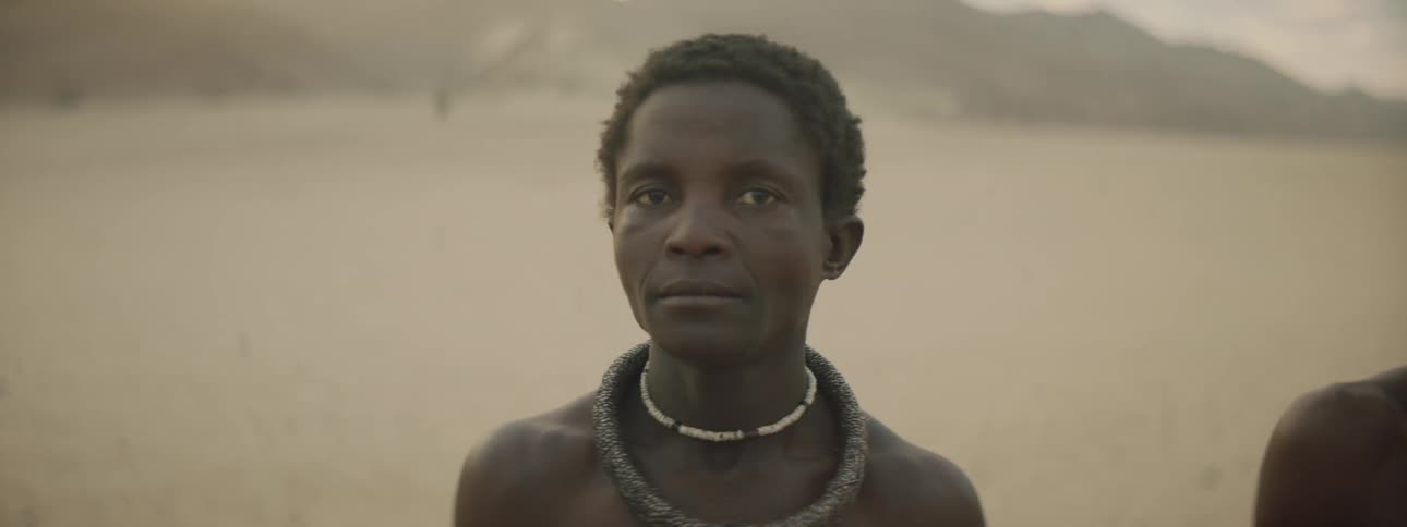 Men stand on bare chested on sand near mountains wearing traditional necklaces