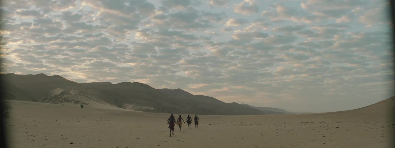 Members in a tribe go running across the desert carrying arrows
