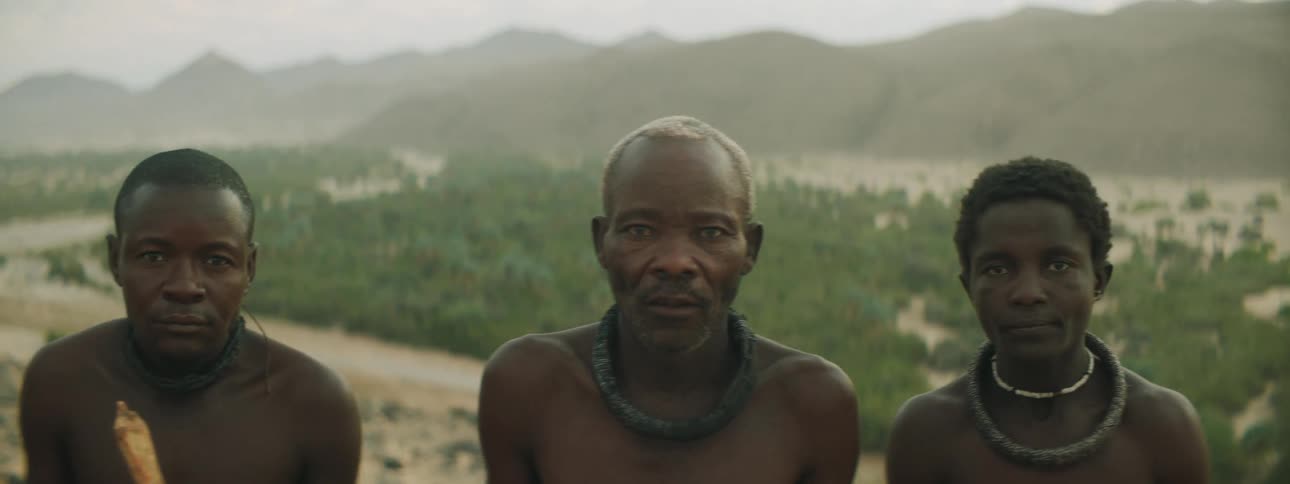 Three men with bare chests and wearing necklaces stand outside with hills in the background