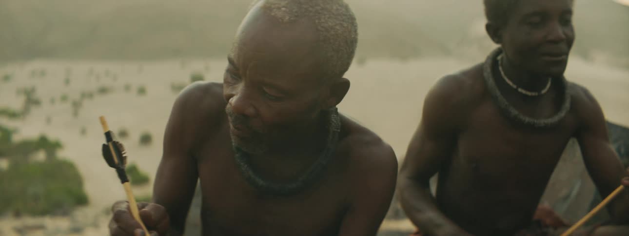 A group of middle aged tribal men examine arrows while sitting on a stone outdoors