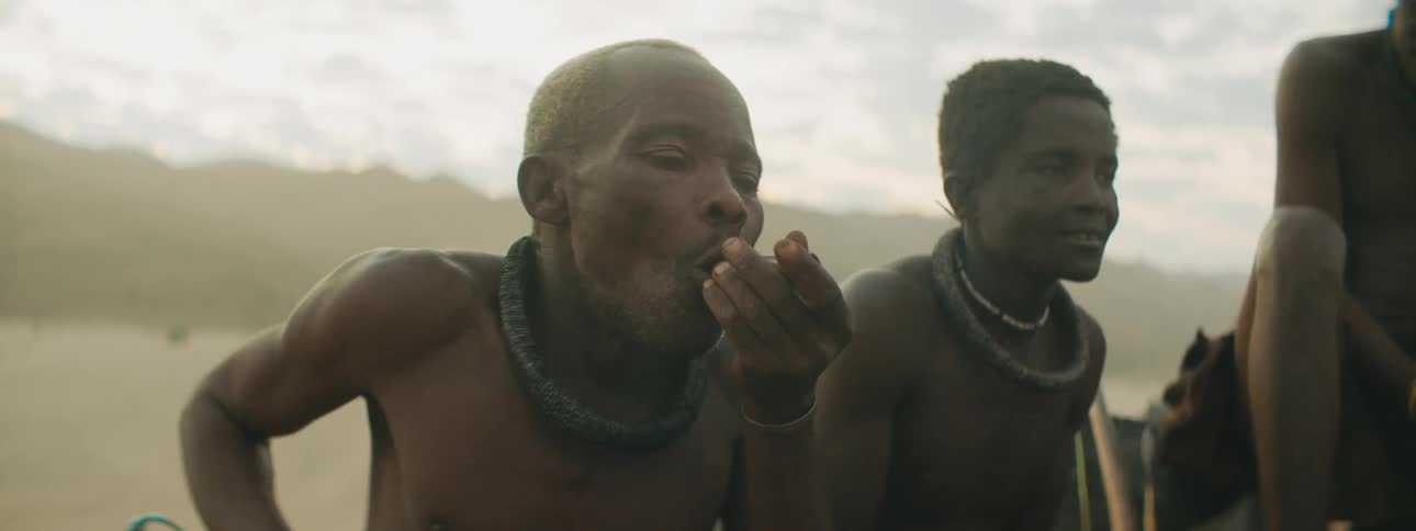 Men in traditional garb sit backed by desert and mountain under cloudy sky smoking