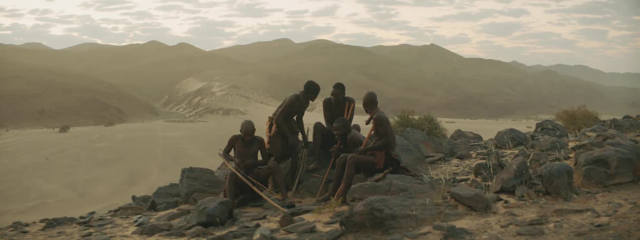 Men sits on rocks working with long staffs backed by desert and misty mountains