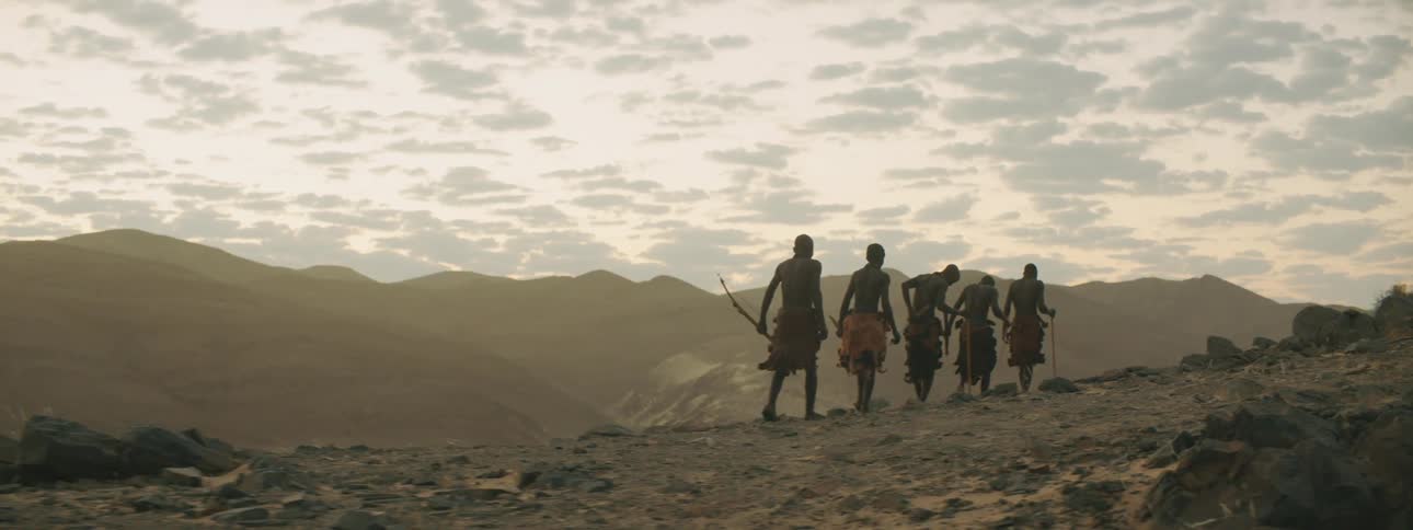 Men walking in a desert with a mountain range the sky and clouds in the background