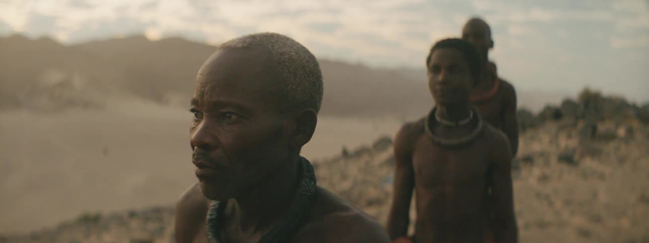 A group of black tribal men stand in line atop a hill in the desert and gaze into the distance