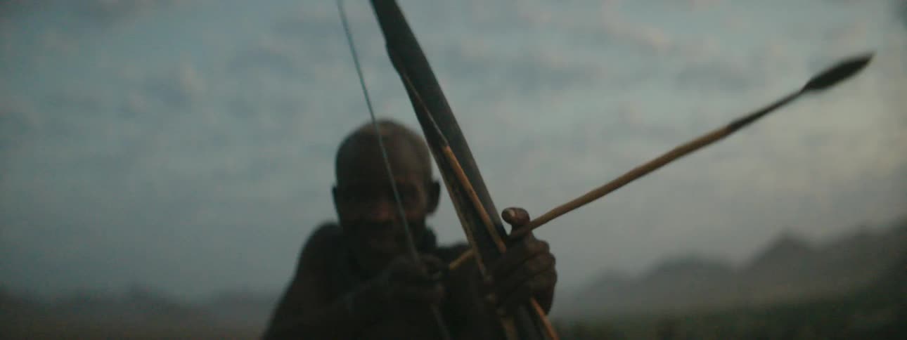 A black tribal man standing outdoors in the evening aims and slowly draws his bow