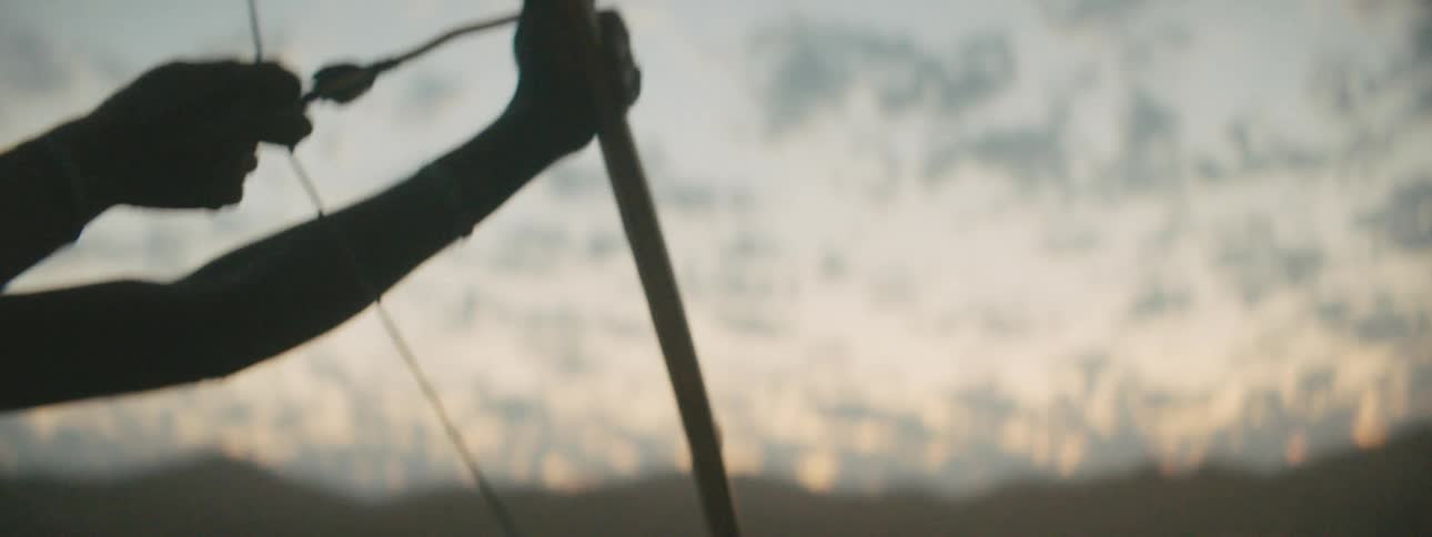 A shirtless man aims and draws a primitive bow while standing outdoors in the evening