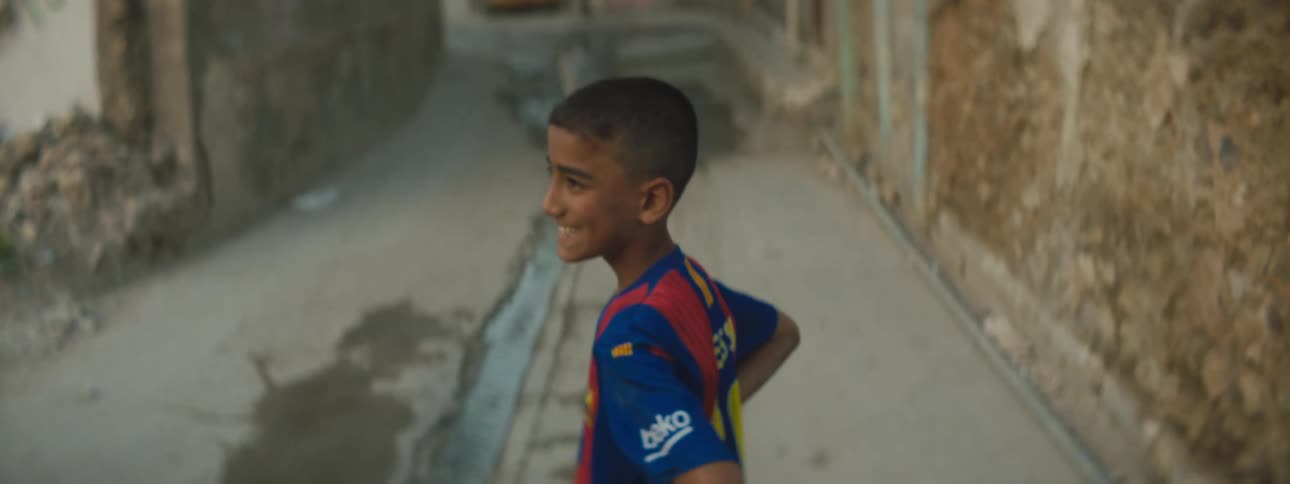 Boy in sports jersey stands in middle of road near stone wall smiling with hands on hips