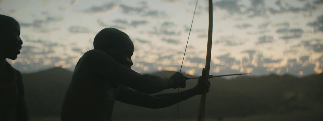 A shirtless man attempts to aim and draw a primitive bow while standing outdoors with another man in a grassland in the evening