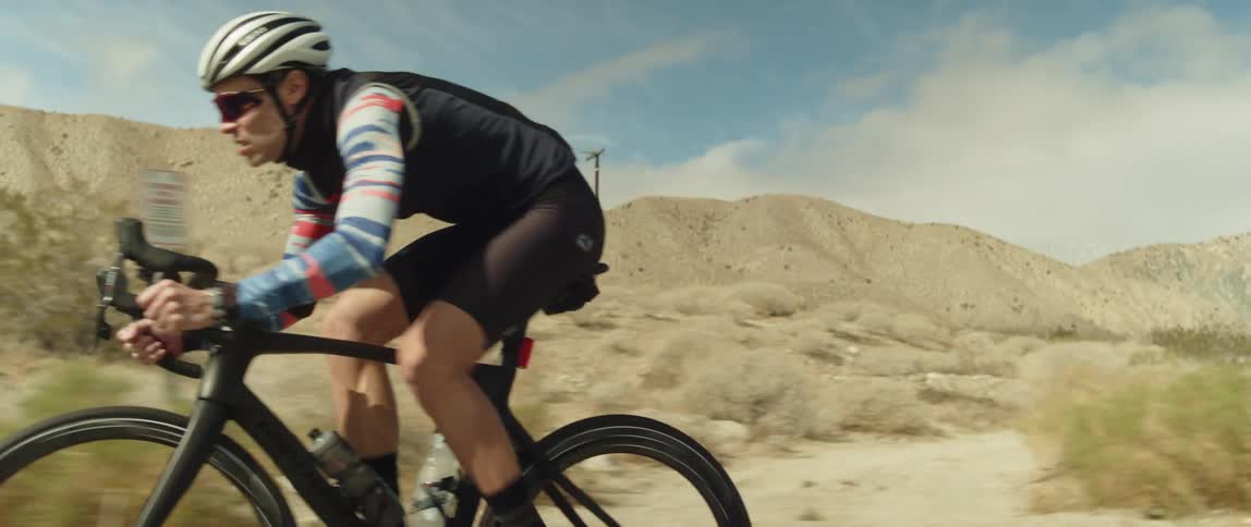 A cyclist pedals a bicycle along a road through a hilly arid countryside during the day