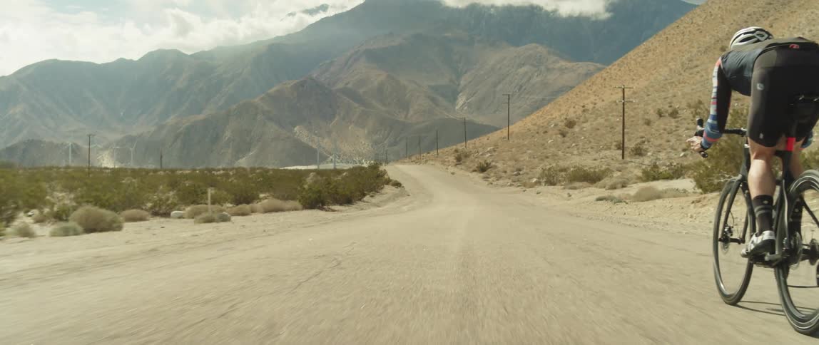 A man wearing a helmet is coasting down a hill on a bike toward mountains