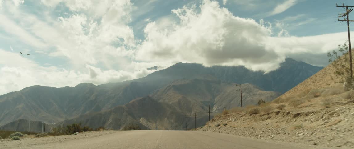 A road through a hilly countryside leading towards a mountain ridge looming in the distance during the day