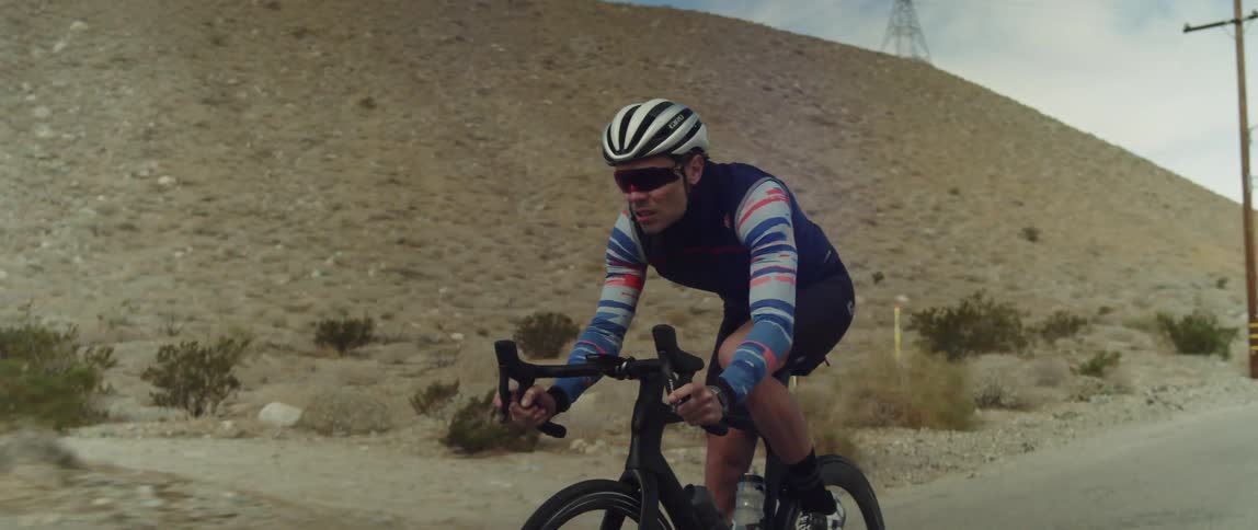 Cyclist rides bicycles along highway past scrub and a hill
