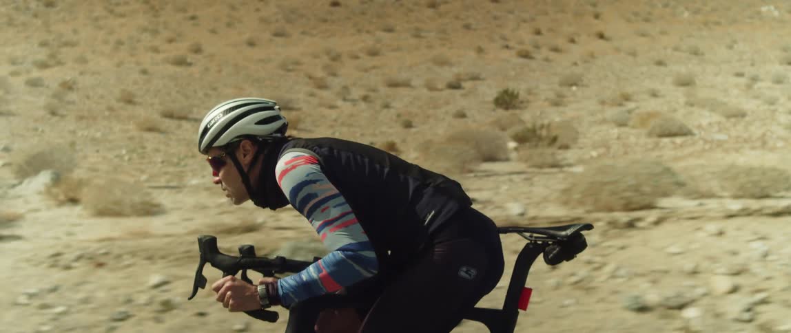 A cyclist ducks down to make himself more aerodynamic while cycling through an arid countryside during the day
