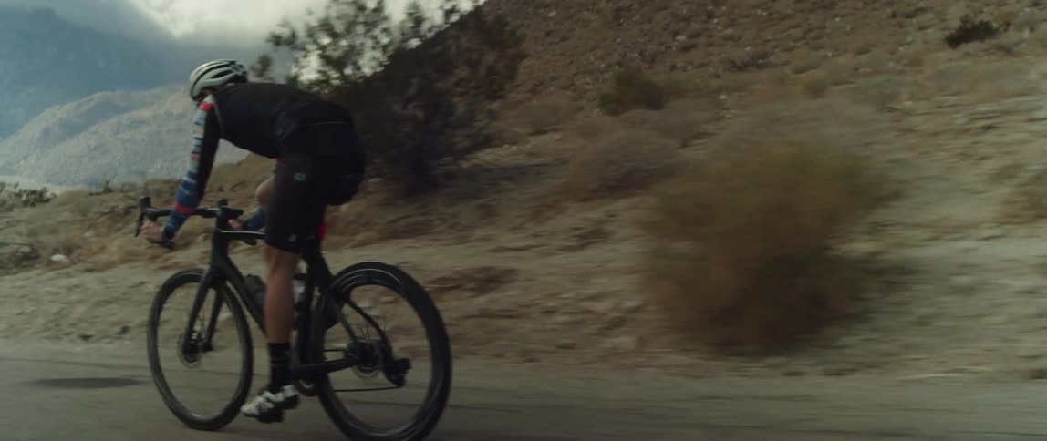 A man wearing a bicycle helmet and sunglasses is riding a bike down a hill near mountains