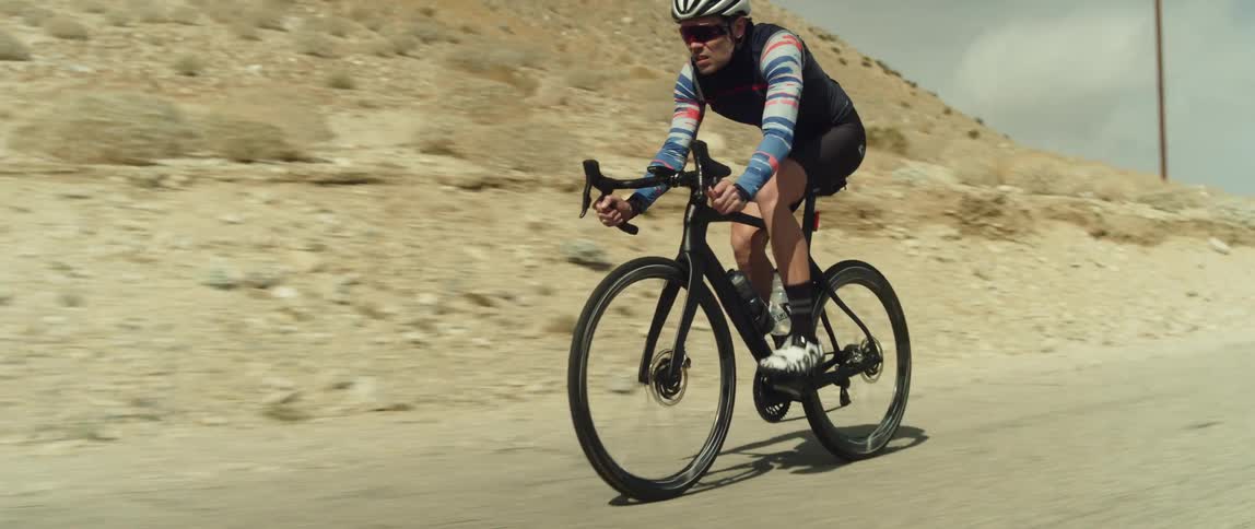 A man rides a bicycle along a road through an arid countryside during the day