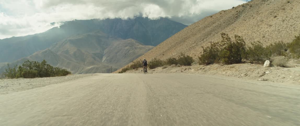 Man wearing a helmet rides a bicycle on a road in the desert close to the mountains