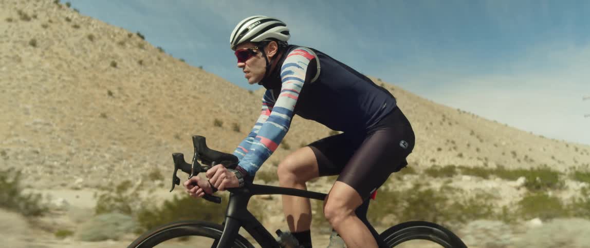 A man wearing a bicycle helmet and sunglasses is riding a bike on a road near mountains
