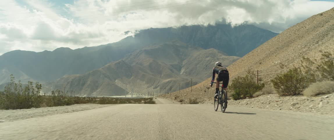 A man wearing a bicycle helmet is riding a bike downhill on a road toward mountains