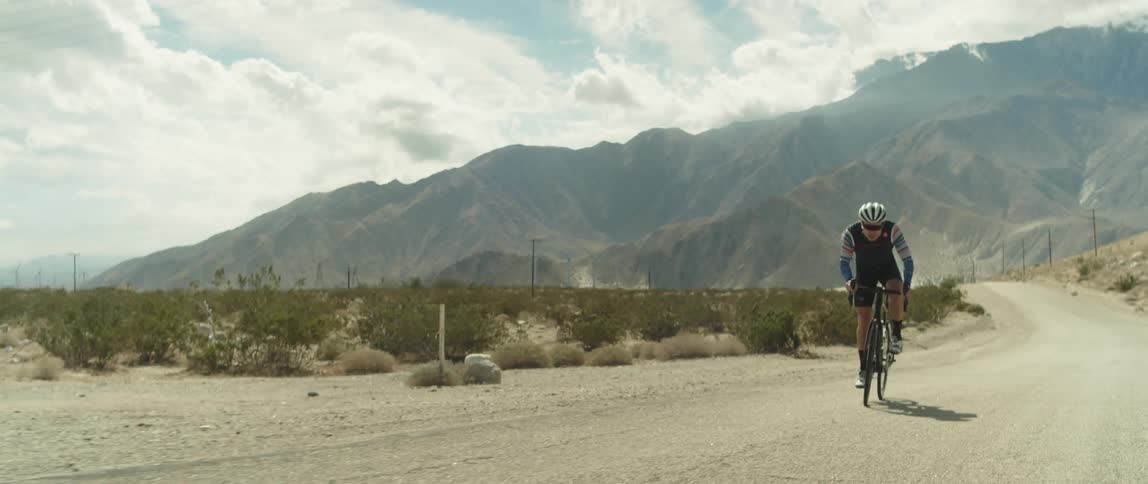 A man wearing a helmet is riding a bike on a dirt road and there are mountains behind him