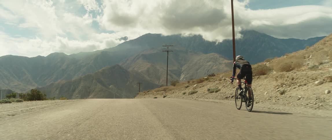 A man wearing a helmet is coasting down a hill on a bike toward mountains