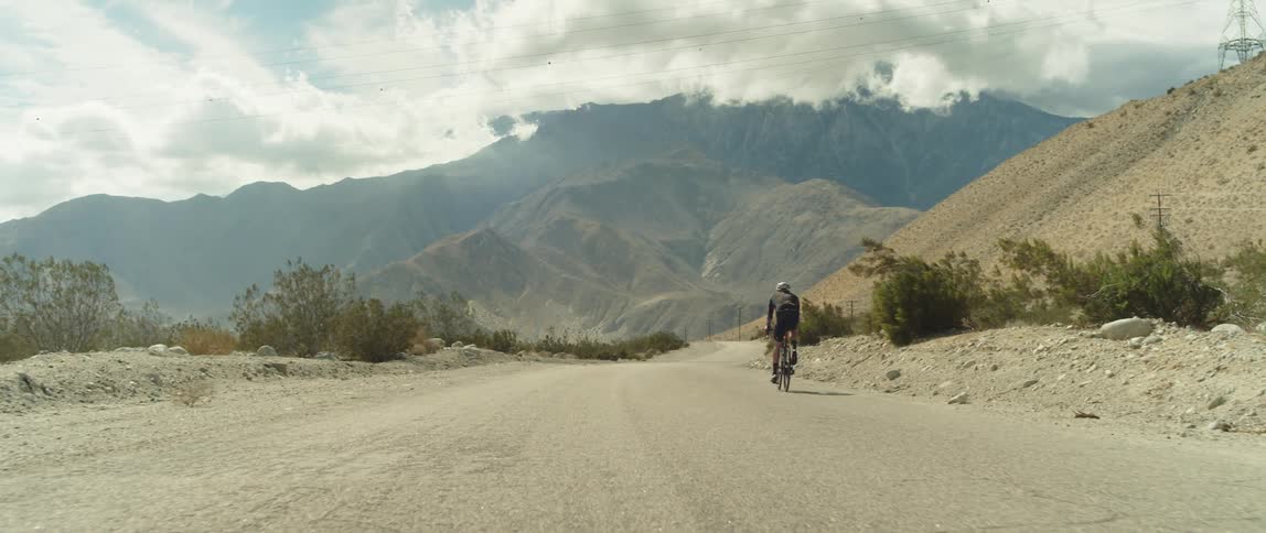 A man wearing a bicycle helmet is riding a bike downhill on a road toward mountains