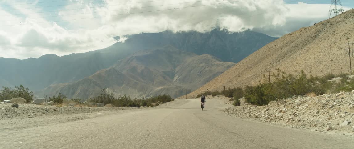 A man is riding a bike on a dirt road near mountains and there are mountains ahead under a cloudy sky