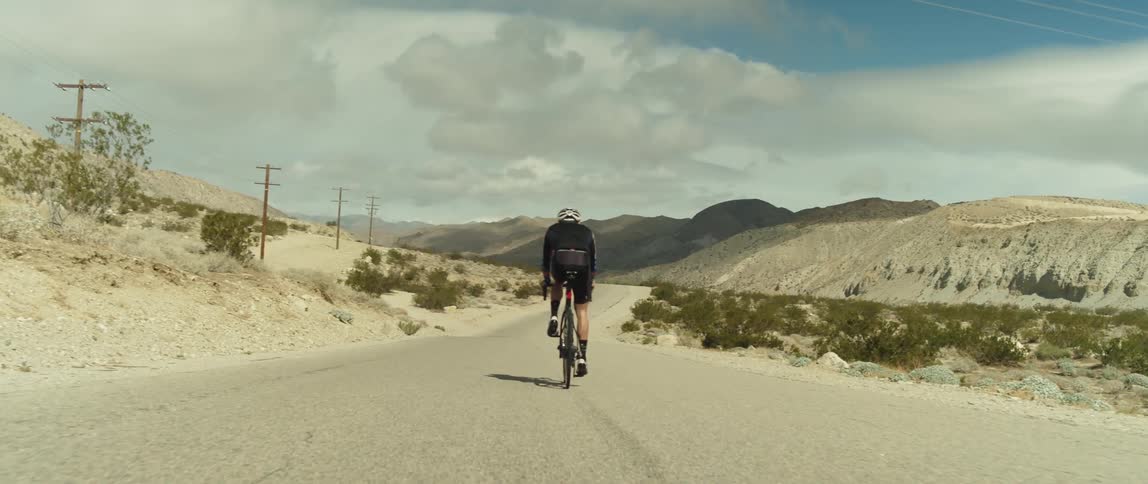 A man wearing a bicycle helmet is riding a bike on a road near mountains under a cloudy sky