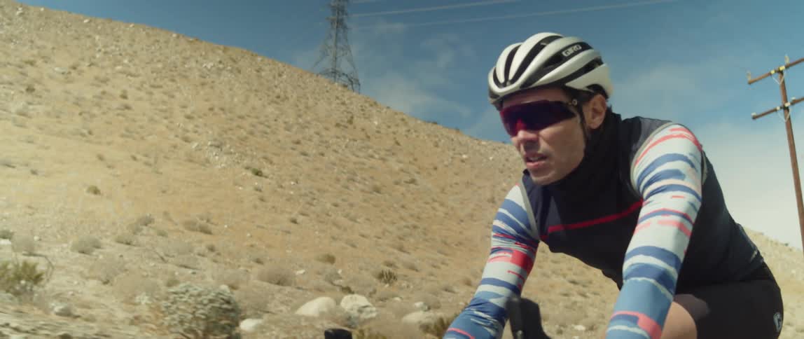 A man wearing a helmet and sunglasses is riding a bike past a utility tower