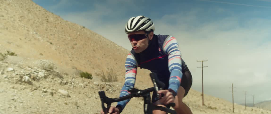 A man wearing a bicycle helmet and sunglasses is riding a bike on a road near mountains