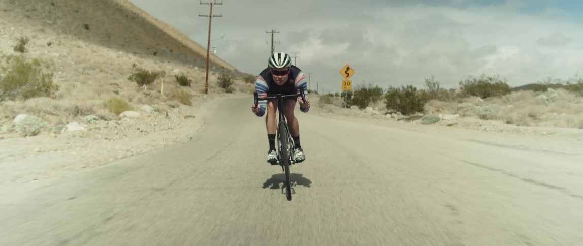 A man wearing a helmet and sunglasses is coasting on a bike under a cloudy sky