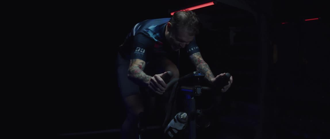Tattooed man pedaling on a static bicycle inside a dimly lit room