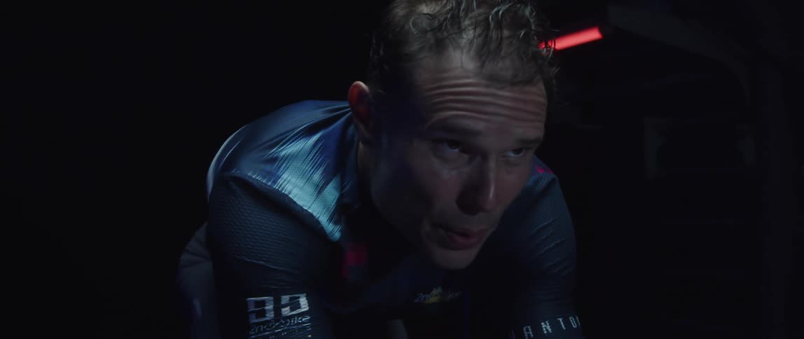 Tattooed man pedaling on a static bicycle inside a dimly lit room