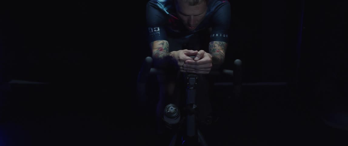 Tattooed man pedaling on a static bicycle inside a dimly lit room