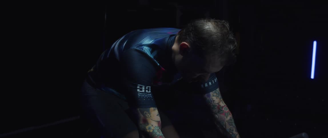 Man with tattoo sleeves pedals on exercise bike in dark room