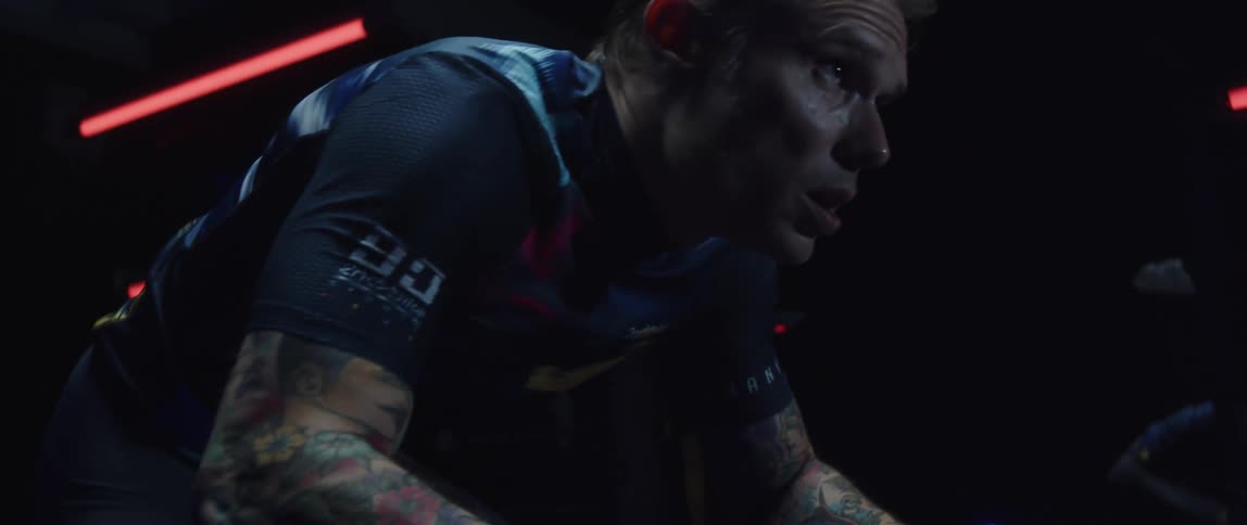 A tattooed man riding a stationary bike in a dark room