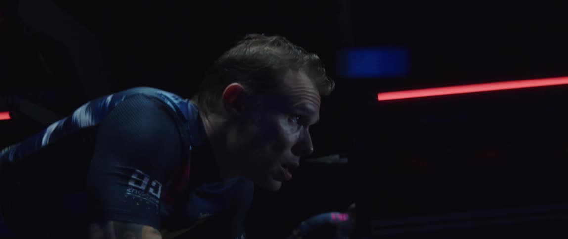 Tattooed man pedaling on a static bicycle inside a dimly lit room