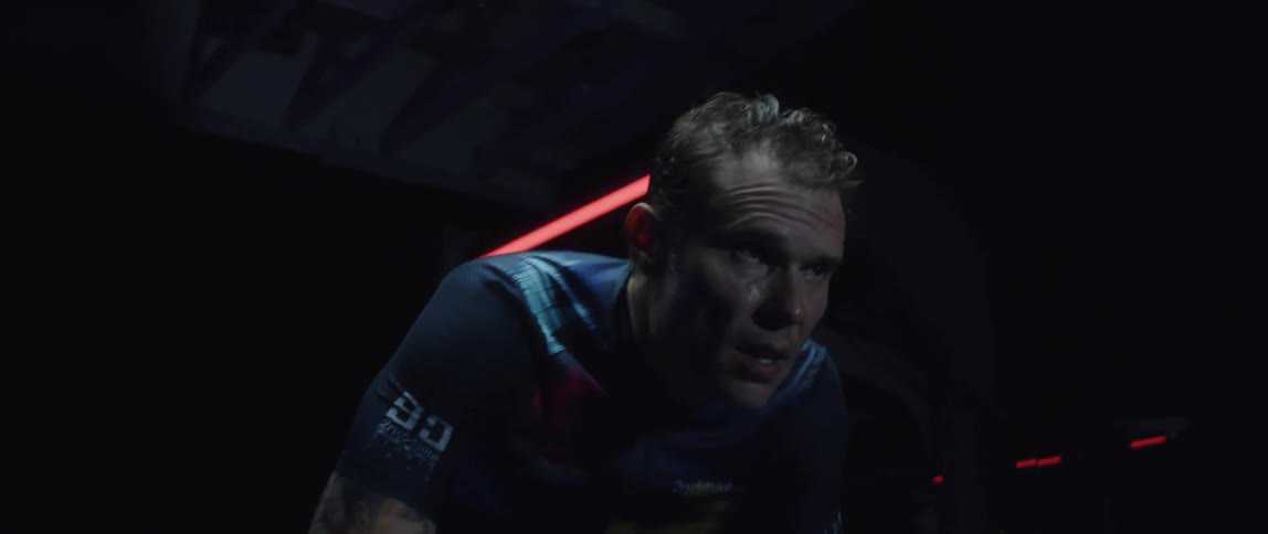 Tattooed man pedaling on a static bicycle inside a dimly lit room