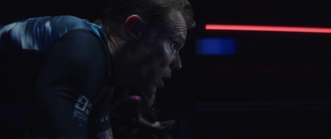 Focused sweaty man heavy breathing while riding a stationary bike in a dark gym room