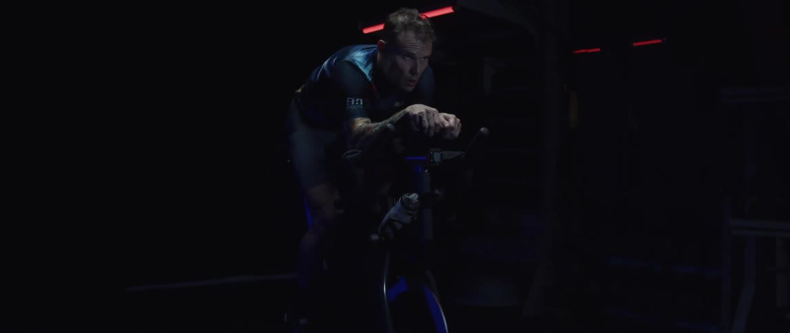 Twisting footage of a man pedaling on a static bicycle inside a dimly lit room against a dark background