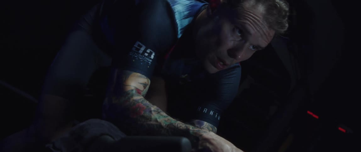 A tattooed man riding a stationary bike in a dimly lit room