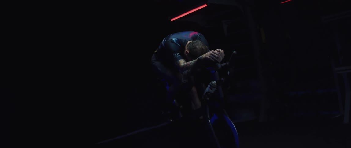 An exhausted man rests atop an exercise bike in a dark room