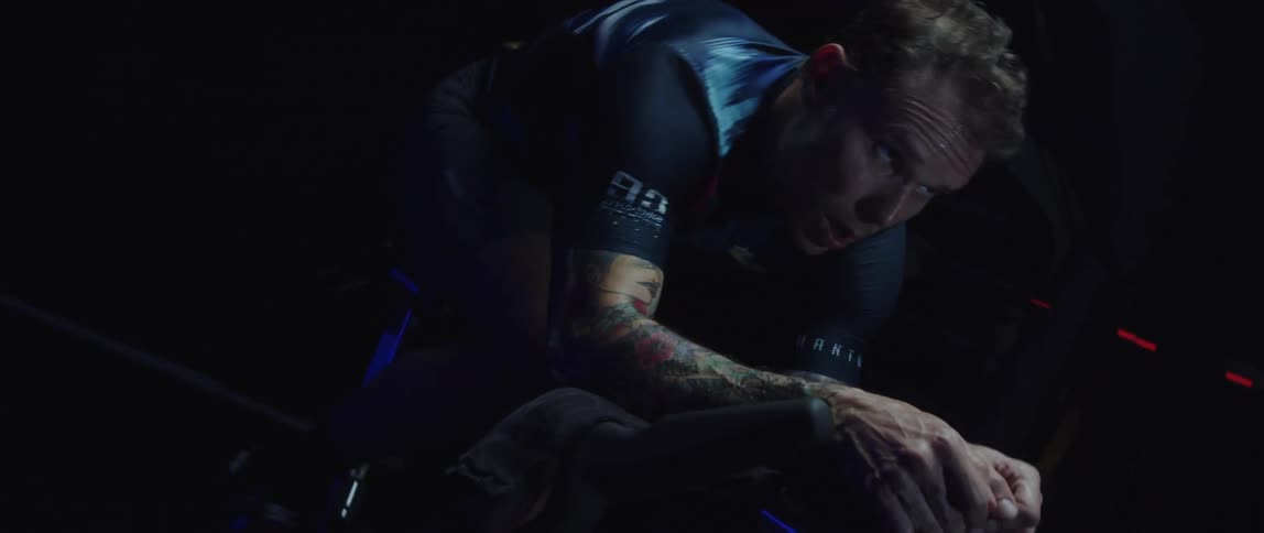 Twisting footage of a man pedaling on a static bicycle inside a dimly lit room against a dark background