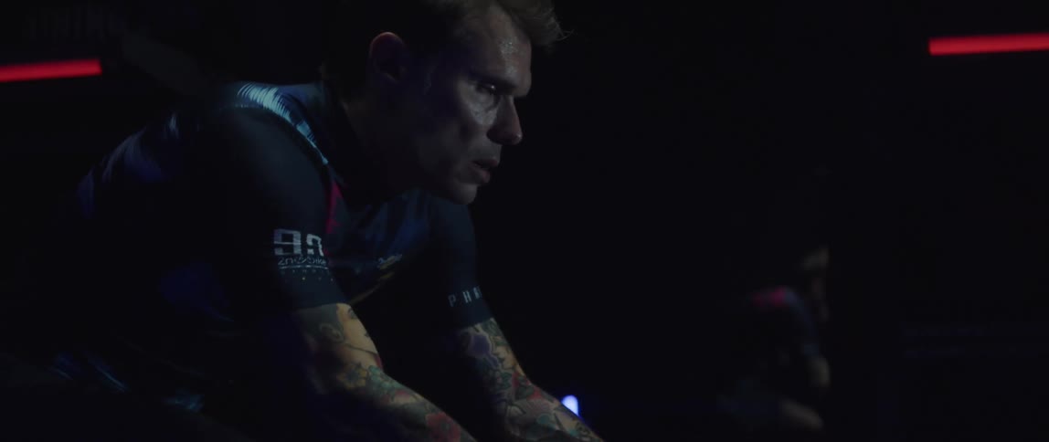 Tattooed man pedaling on a static bicycle inside a dimly lit room